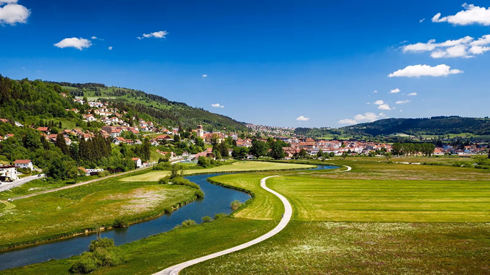 Curved river flowing through green fields toward a village with houses and hills under a blue sky.