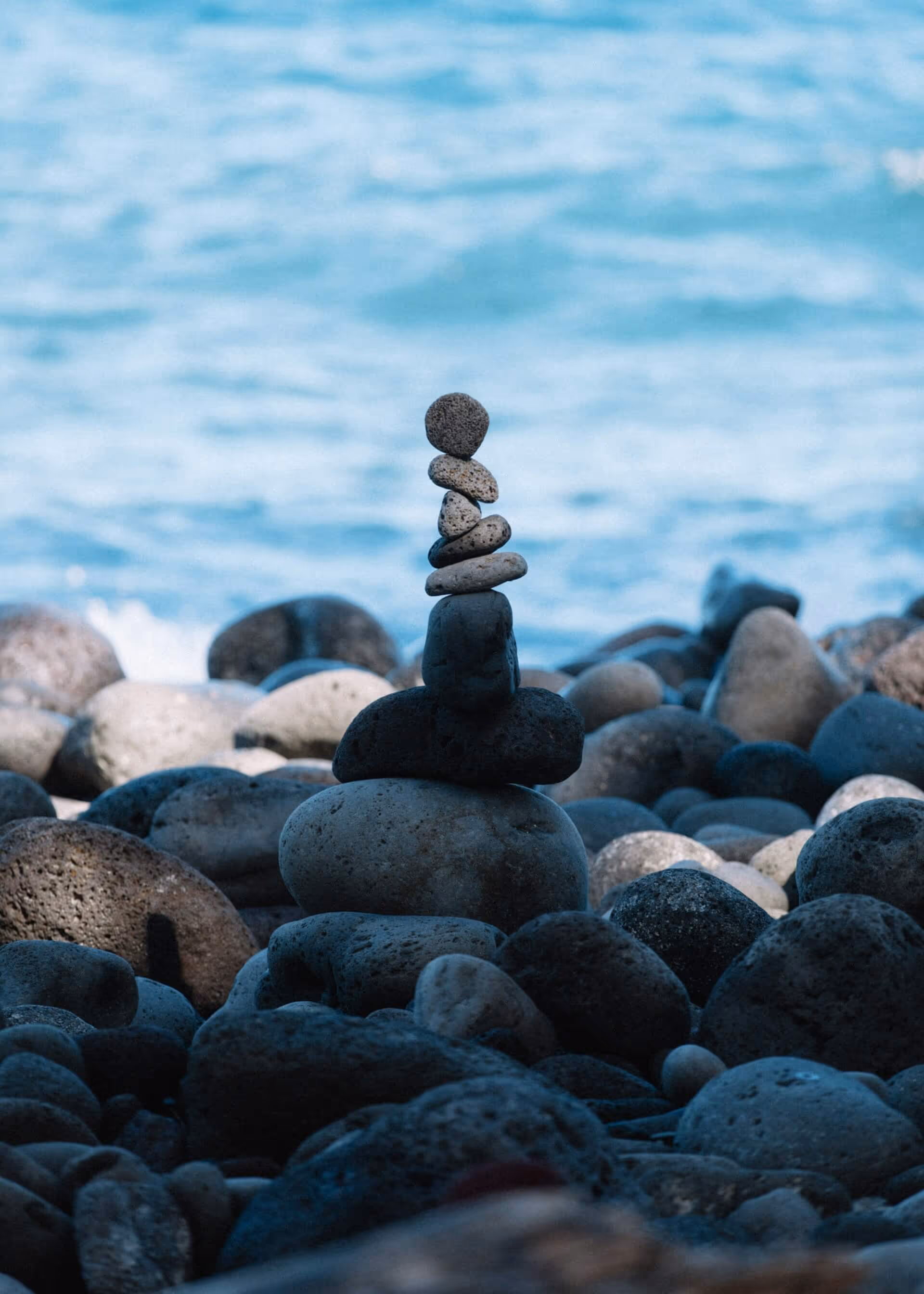Stack of balanced stones on a rocky shore with blurred ocean water in the background.