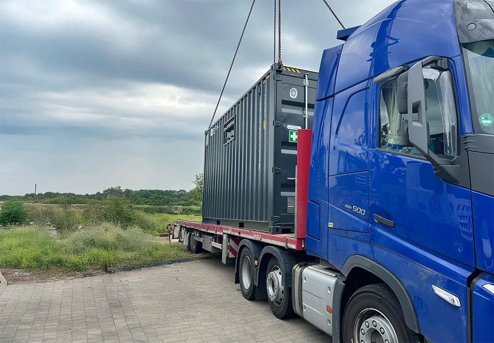 Large blue truck with a flatbed carrying a black shipping container being lifted by chains while a man stands nearby on a paved surface by grassy fields under a cloudy sky.
