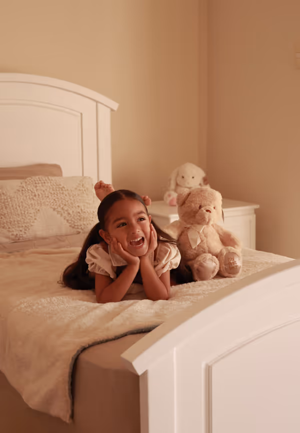 Niña sonriente acostada boca abajo en la cama con un osito de peluche al lado en una habitación iluminada con luz cálida.