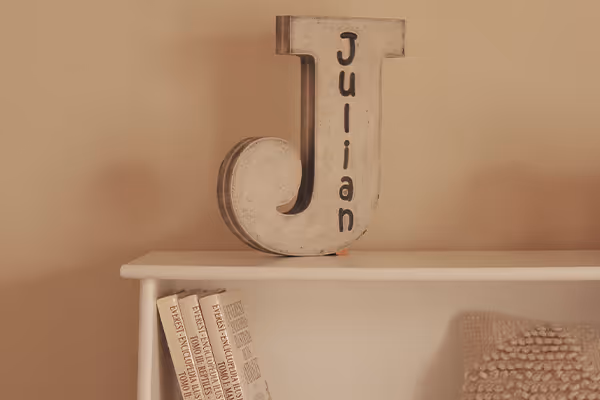 Letrero decorativo grande en forma de J con el nombre Julian, sobre una repisa blanca con libros y una manta texturizada.