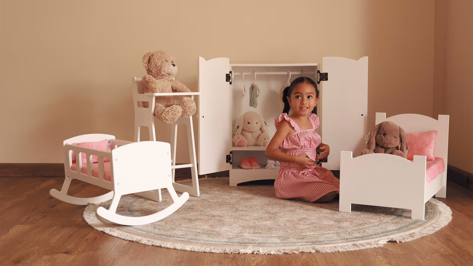 Niña sentada en un tapete redondo junto a muebles de juguete blancos y peluches en una habitación con piso de madera.
