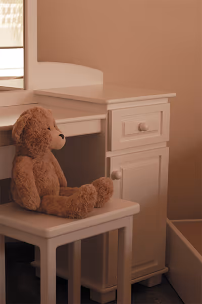 Un oso de peluche marrón sentado en una silla blanca frente a un tocador blanco con cajones.