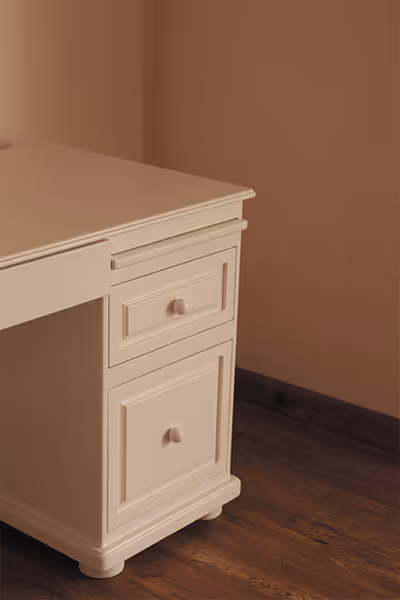 Detalle lateral de un escritorio blanco con dos cajones, sobre un suelo de madera y fondo de pared blanca.