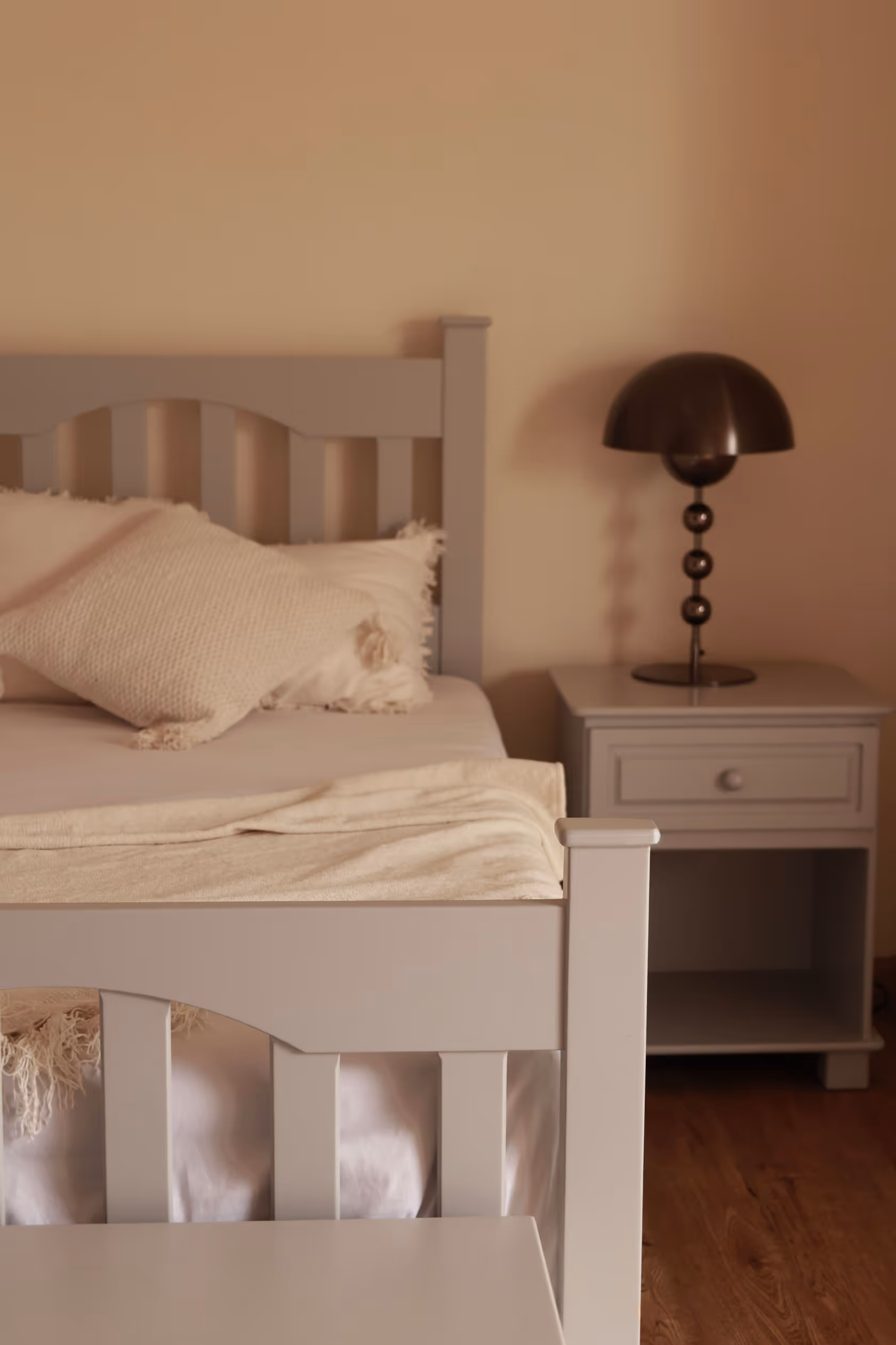 Cama gris con almohada y manta beige, junto a una mesa de noche del mismo color con lámpara negra.