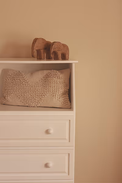 Cojín texturizado en tonos beige sobre una cómoda blanca con dos elefantes decorativos de madera encima.