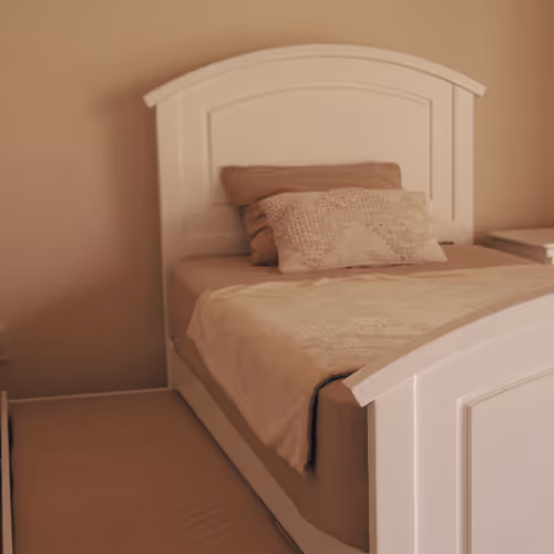 Cama individual con cabecera y pie de cama blancos, sábanas y almohadas en tonos beige en un dormitorio.