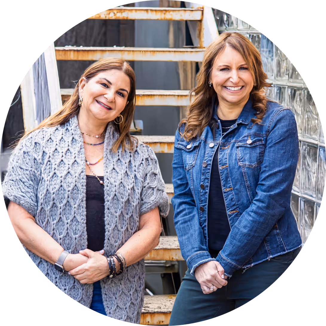 Amy and Debbie smiling and posing in front of a rusted metal staircase; one wearing a grey knitted shawl and the other a blue denim jacket.