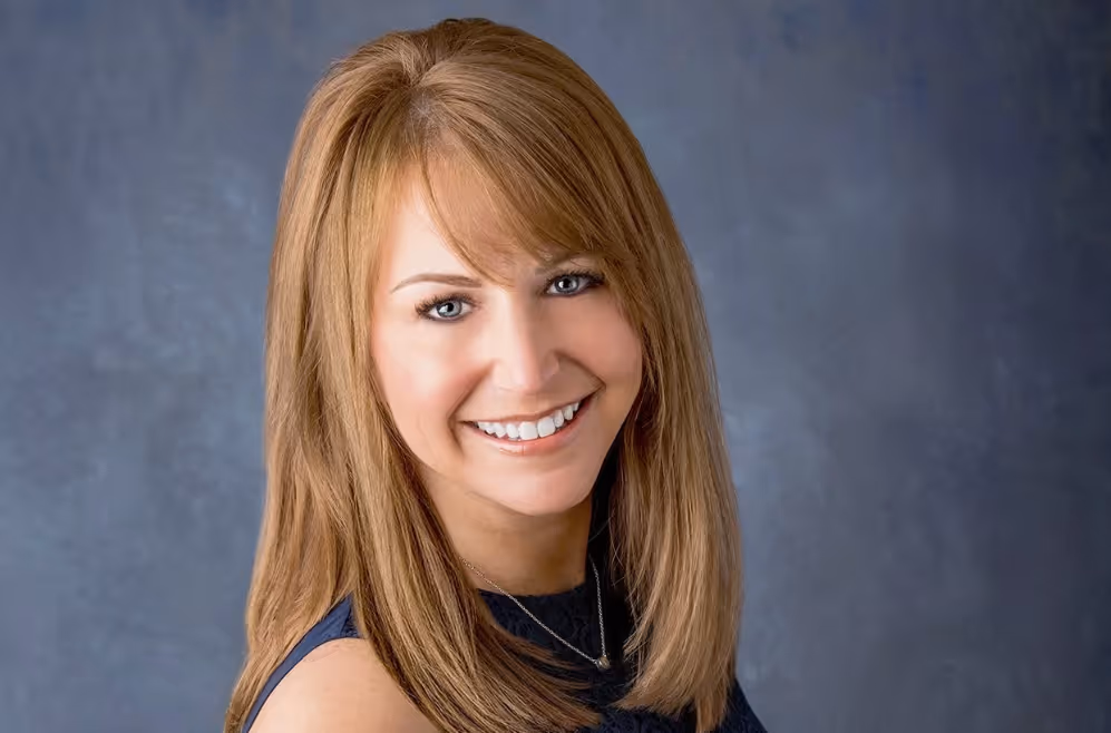 Smiling Debbie Kanter with straight light brown hair wearing a sleeveless dark top against a gray background.