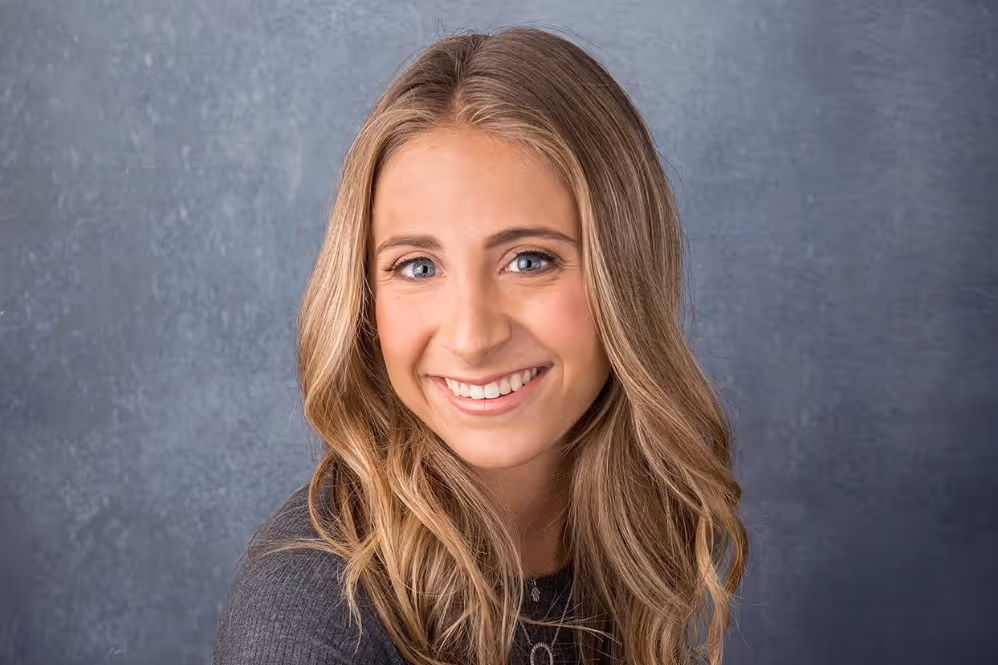 Smiling young woman with long blonde hair wearing a gray top against a textured gray background.