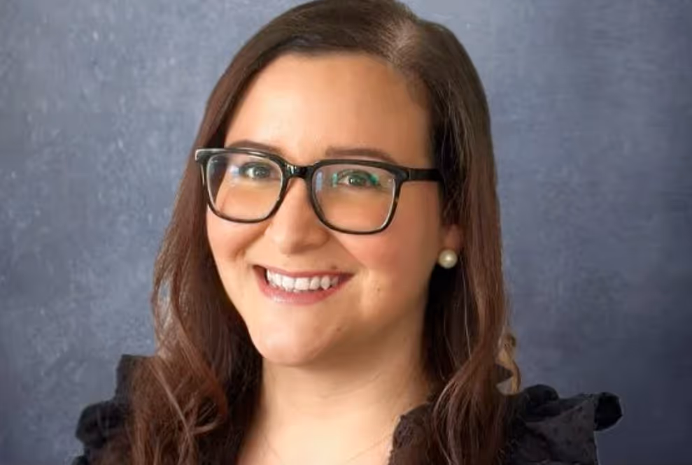 Portrait of a smiling woman with long brown hair, glasses, pearl earrings, and a black top against a gray background.