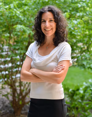 Smiling Laura McDermott with curly dark hair standing outside with her arms crossed, wearing a white top and black skirt.