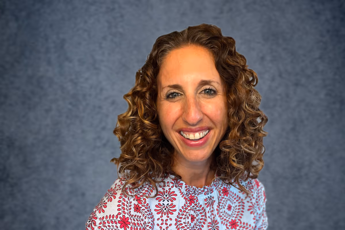 Smiling Leslie Randolph with curly hair wearing a white shirt with red floral patterns against a gray background.