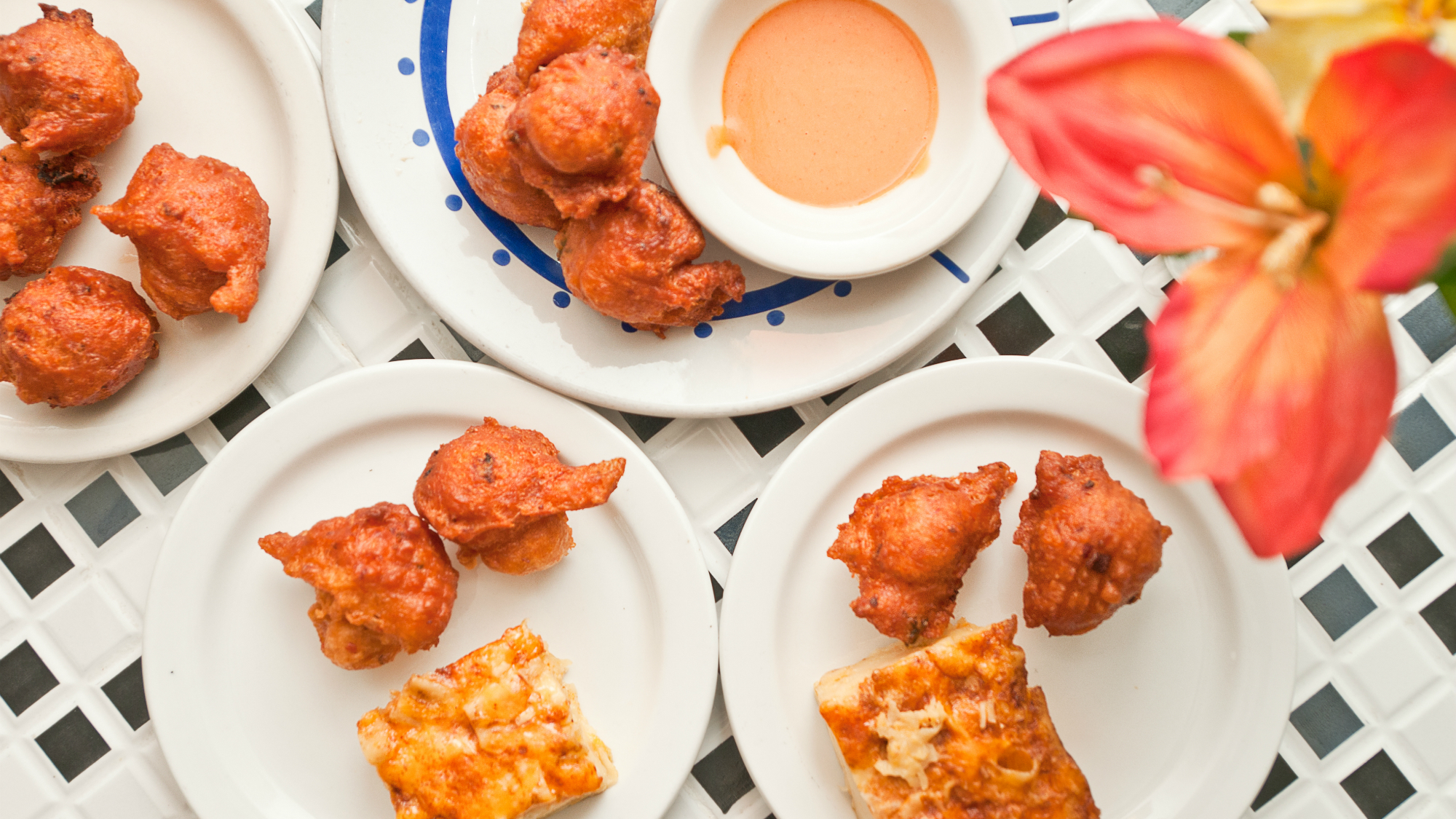 Plates with fried dough fritters, a slice of cheesy pizza, and a bowl of dipping sauce on a white lattice table next to a red flower.