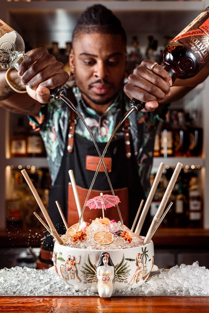 Bartender pouring two bottles of liquor into a large decorative bowl filled with crushed ice and garnished with flowers, a dried citrus slice, and a cocktail umbrella.