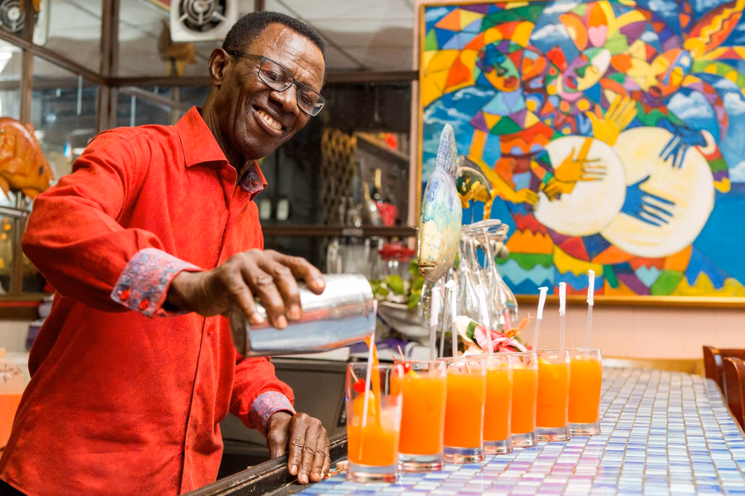 Smiling man in a red shirt pouring orange drinks into a row of glasses on a tiled bar counter in Nassau Bahamas.