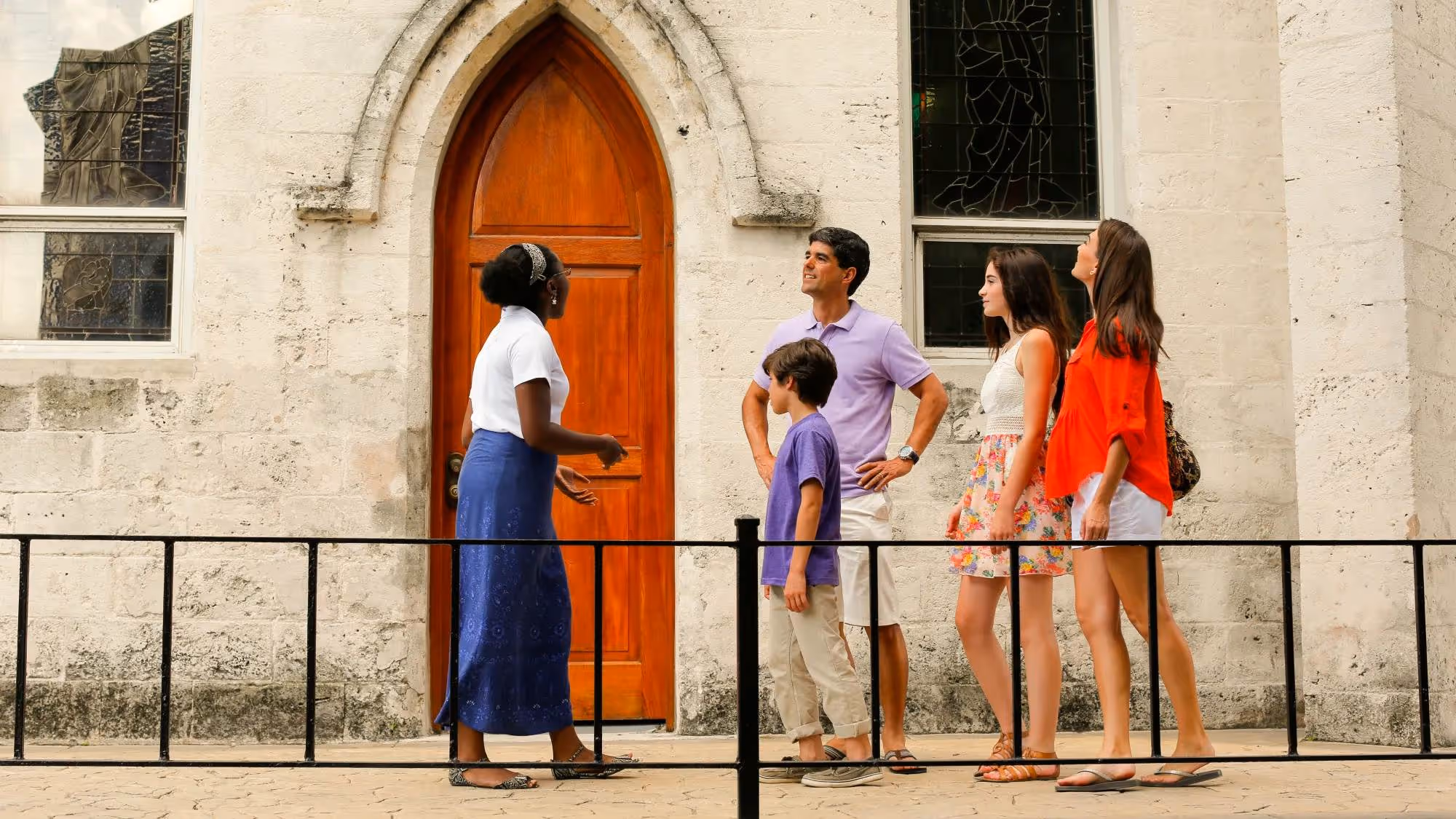 Tour guide on Tru Bahamian Tours Private Tour speaking to a family of four outside a stone building with a wooden arched door and stained-glass windows.