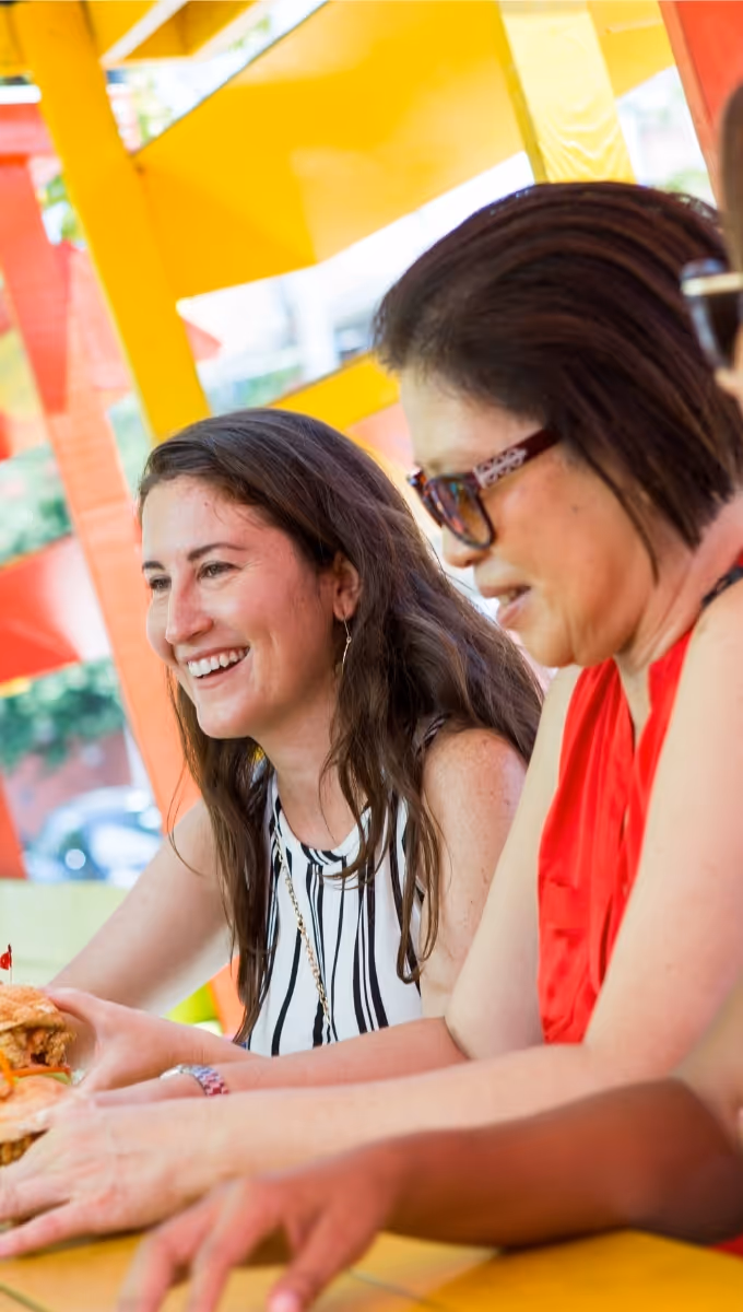 Two women sitting at a table with colorful yellow and red background; one woman is smiling while holding a sandwich in Nassau Bahamas. 