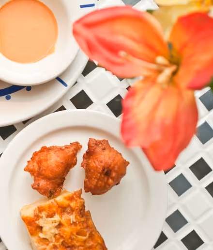 White plate with two fried fritters and a piece of baked pastry on a white lattice table, with an orange flower and a cup of coffee nearby.