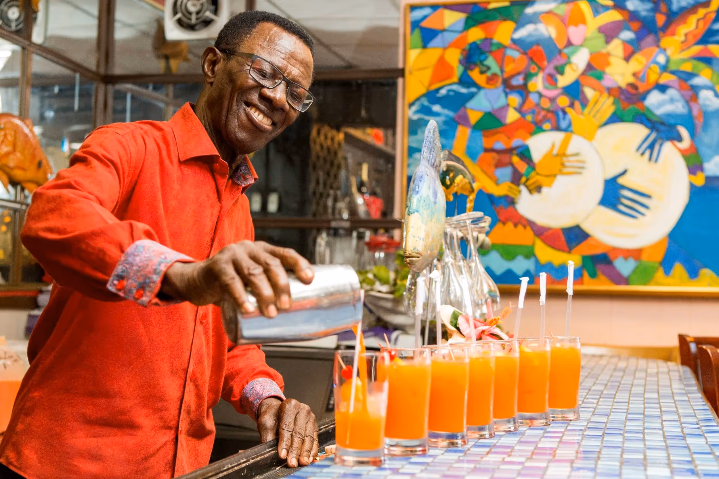 Smiling man in red shirt pouring orange drinks into glasses lined up on a tiled bar counter with colorful abstract painting in background.