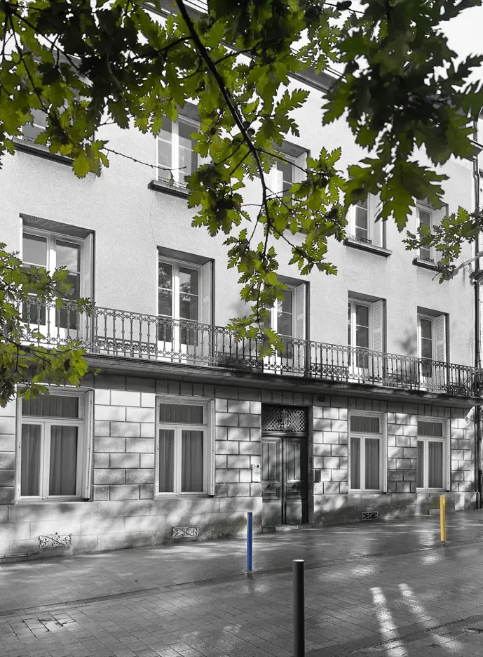 Façade d'un bâtiment avec des fenêtres et une porte, vue partielle d'arbres aux feuilles vertes au premier plan.