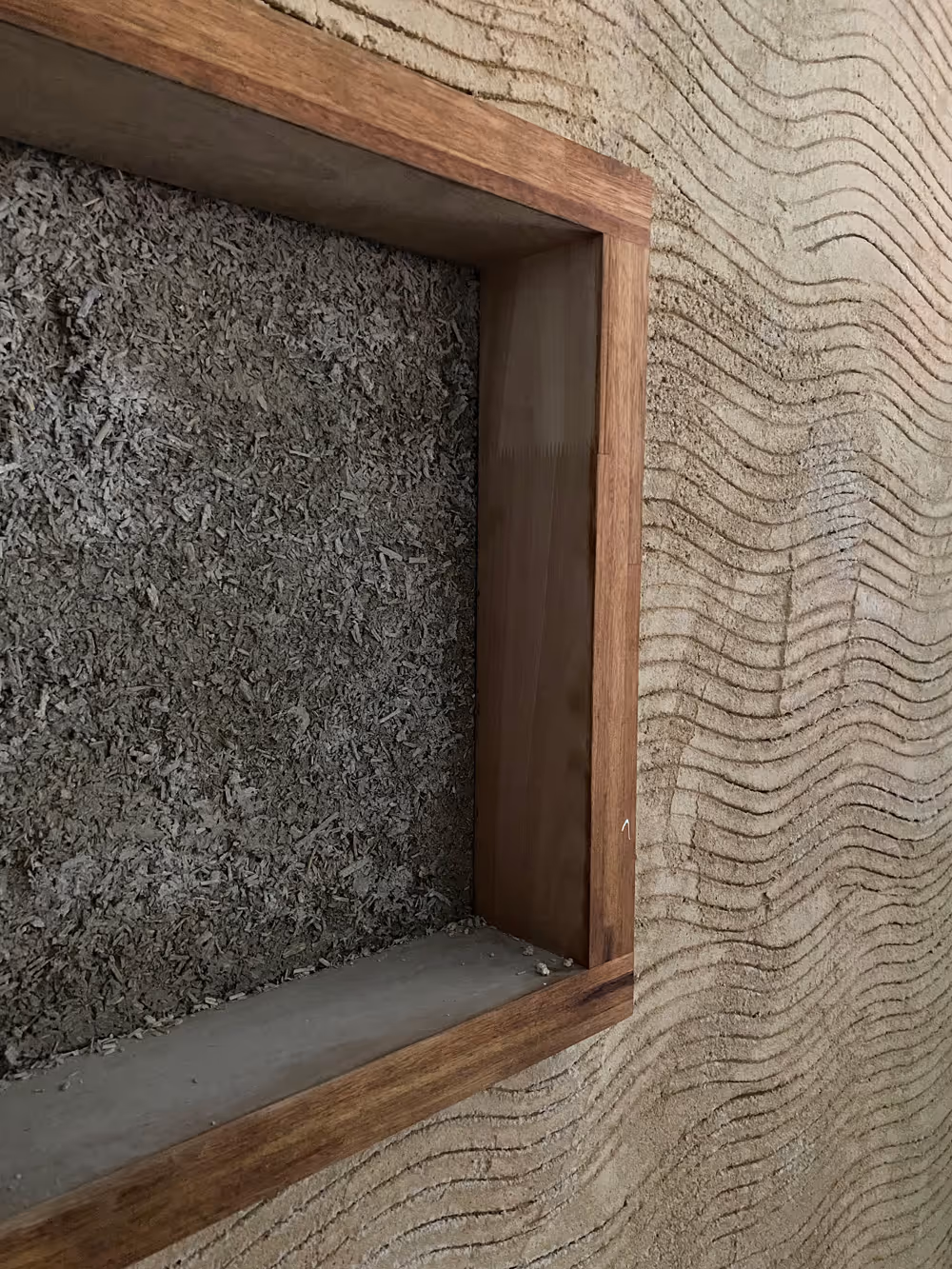 Mur en béton texturé avec motif ondulé et encadrement en bois autour d'une niche remplie de gravier.