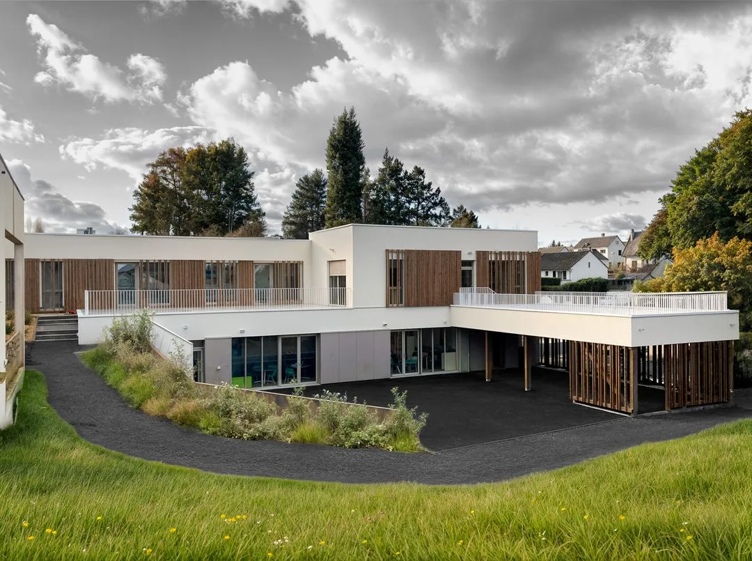 Bâtiment moderne à deux étages avec des panneaux en bois, entouré d'herbe verte et d'arbres sous un ciel nuageux.