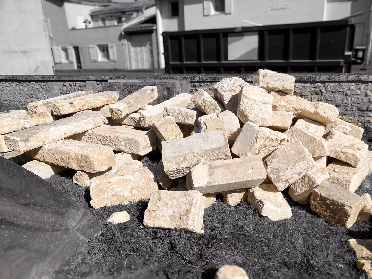 Pile of beige rectangular limestone bricks stacked outdoors on a grassy area with a stone wall and buildings in the background.