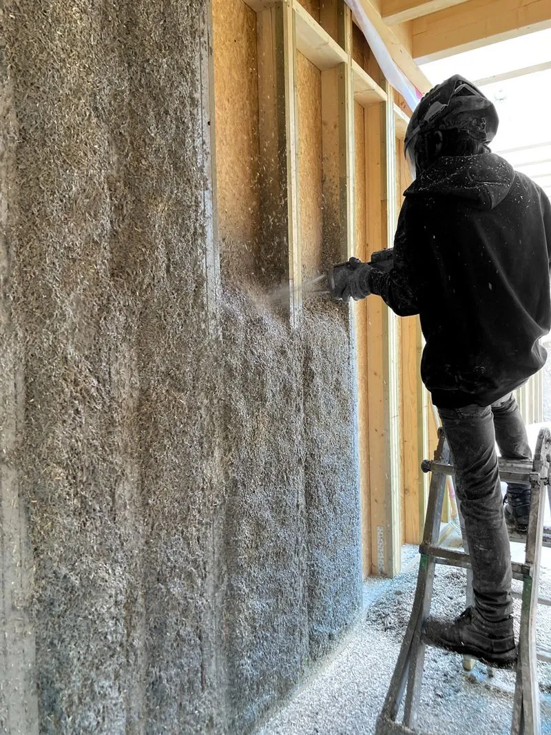 Un ouvrier en tenue noire applique de l'isolant naturel sur une ossature en bois à l'aide d'une machine pulvérisatrice.