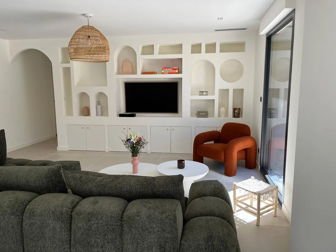 Salon lumineux avec canapé gris, fauteuil orange, tables blanches, meuble TV blanc avec niches décoratives et grande fenêtre vitrée.