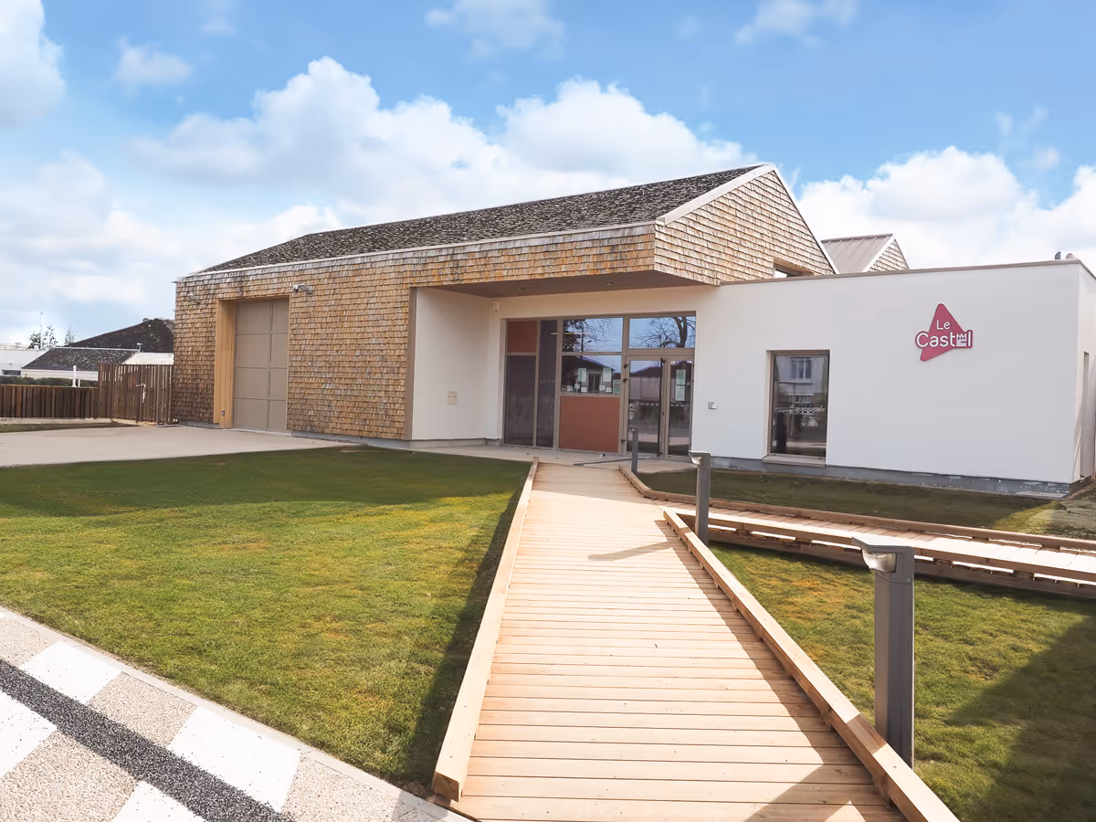 Bâtiment moderne avec bardage en bois et façade blanche, chemin en bois menant à l'entrée sous un ciel bleu avec quelques nuages.