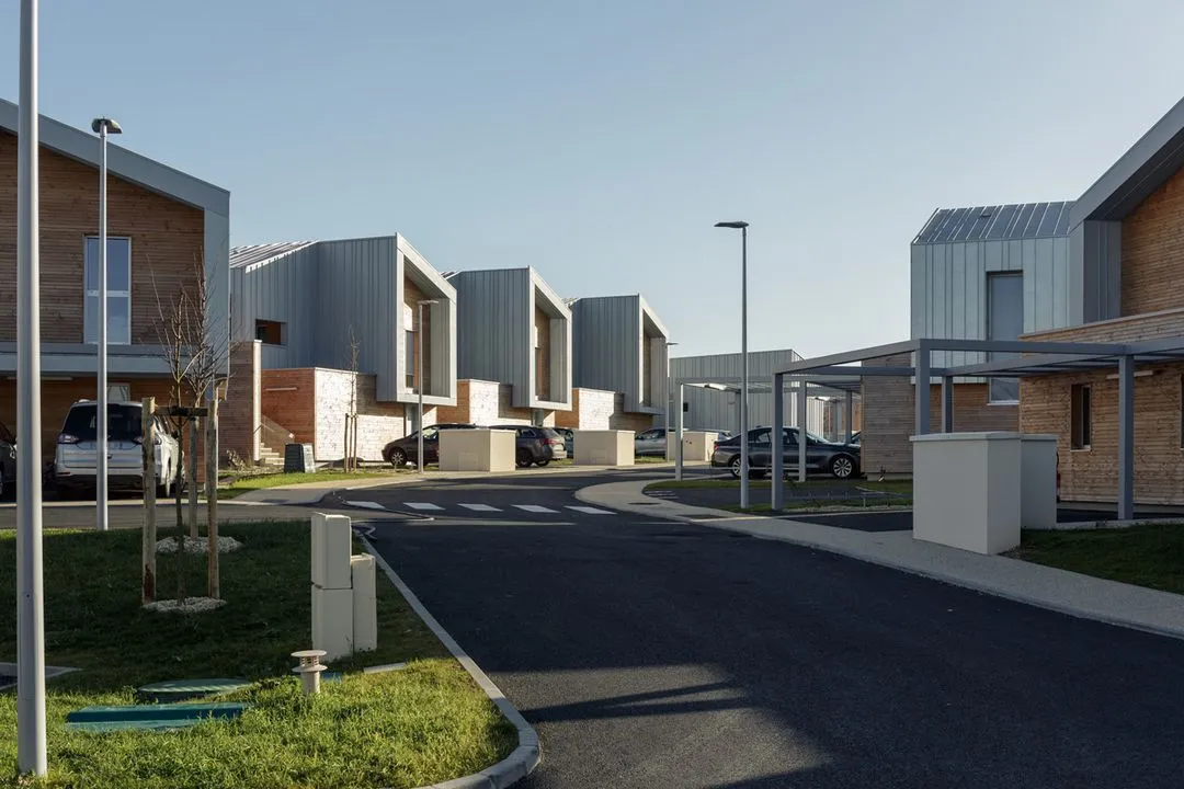 Quartier résidentiel moderne avec des maisons en bois et métal, voitures garées et éclairage urbain sous un ciel clair.