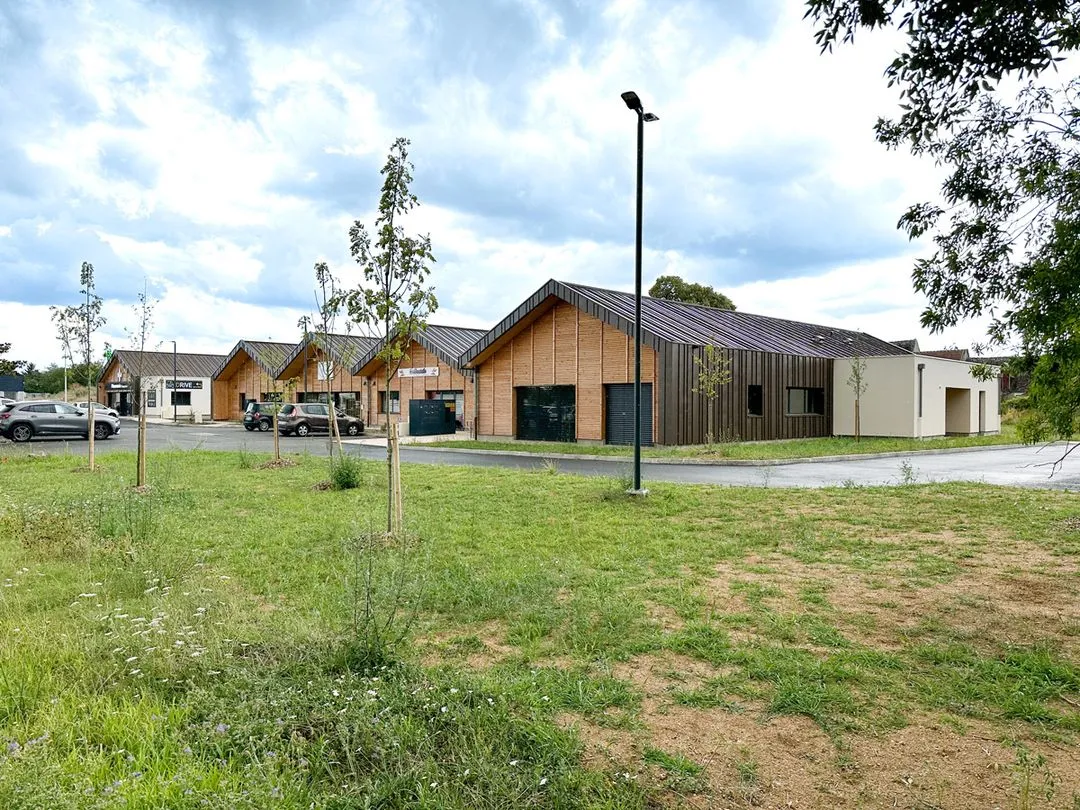 Un bâtiment commercial moderne avec toits en pente, murs en bois et en plâtre, entouré d'arbres et d'une pelouse verte sous un ciel nuageux.