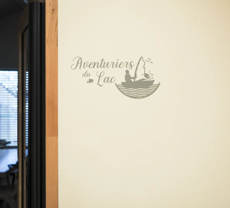 Logo gris sur un mur clair représentant un pêcheur dans un bateau avec des vagues, des oiseaux, un poisson et le texte « Aventuriers du Lac ».