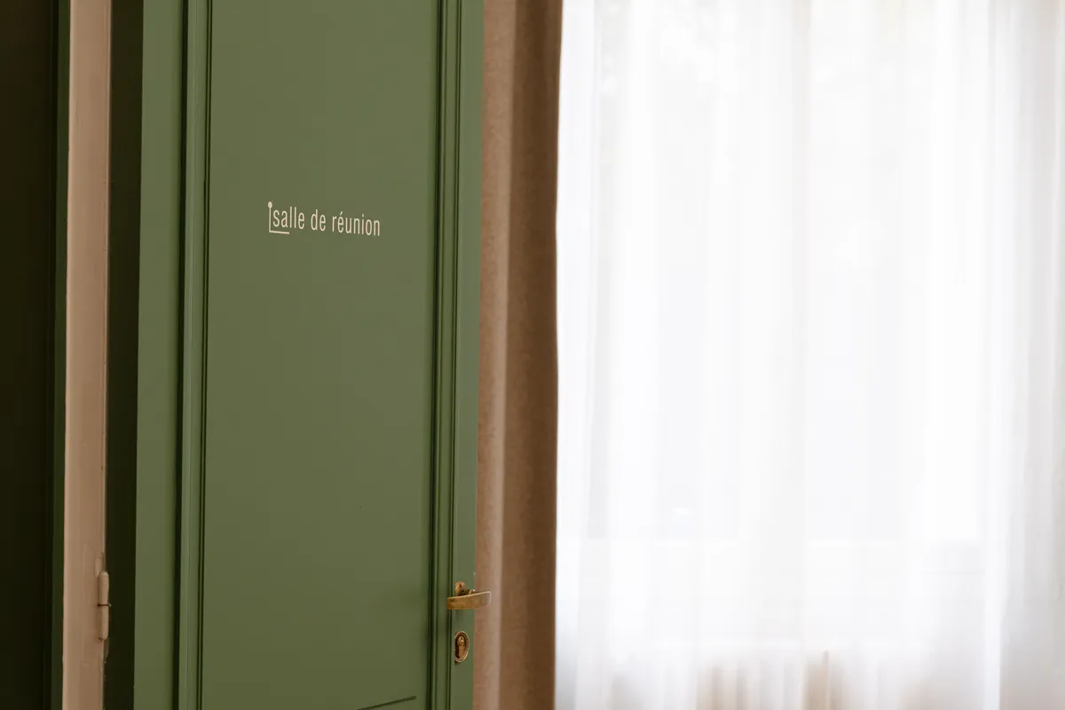 Porte verte avec l'inscription « salle de réunion » sur un mur clair à côté d'une fenêtre avec un rideau blanc.