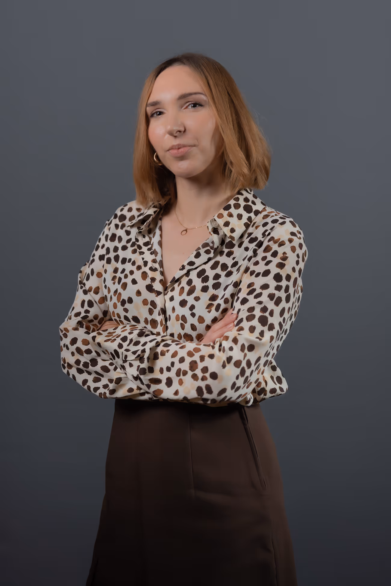 Femme aux cheveux courts roux debout, bras croisés, portant une chemise à motif léopard et une jupe marron devant un fond gris.