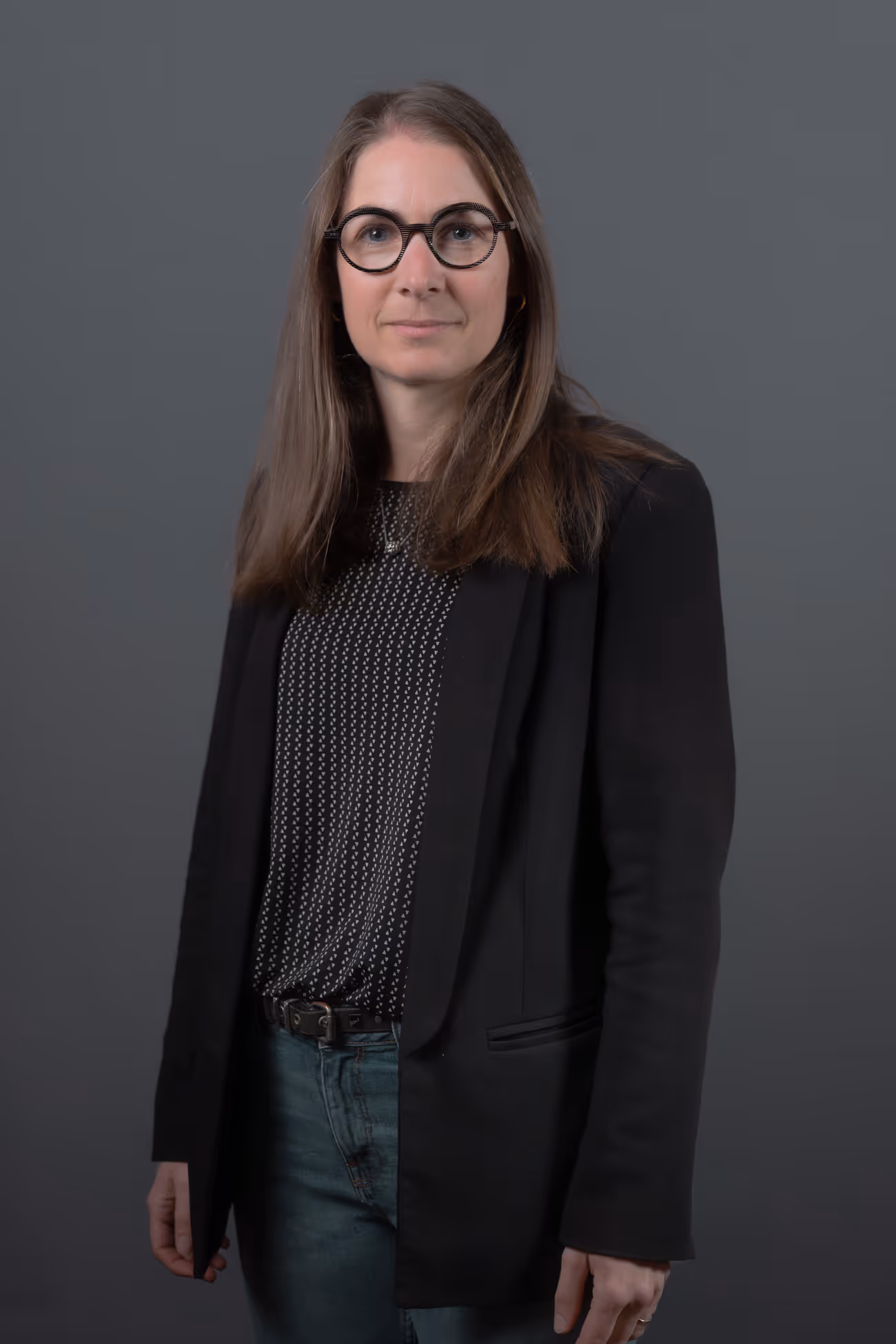 Portrait d'une femme portant des lunettes rondes, un blazer noir, un chemisier à motifs et un jean bleu, sur fond gris.