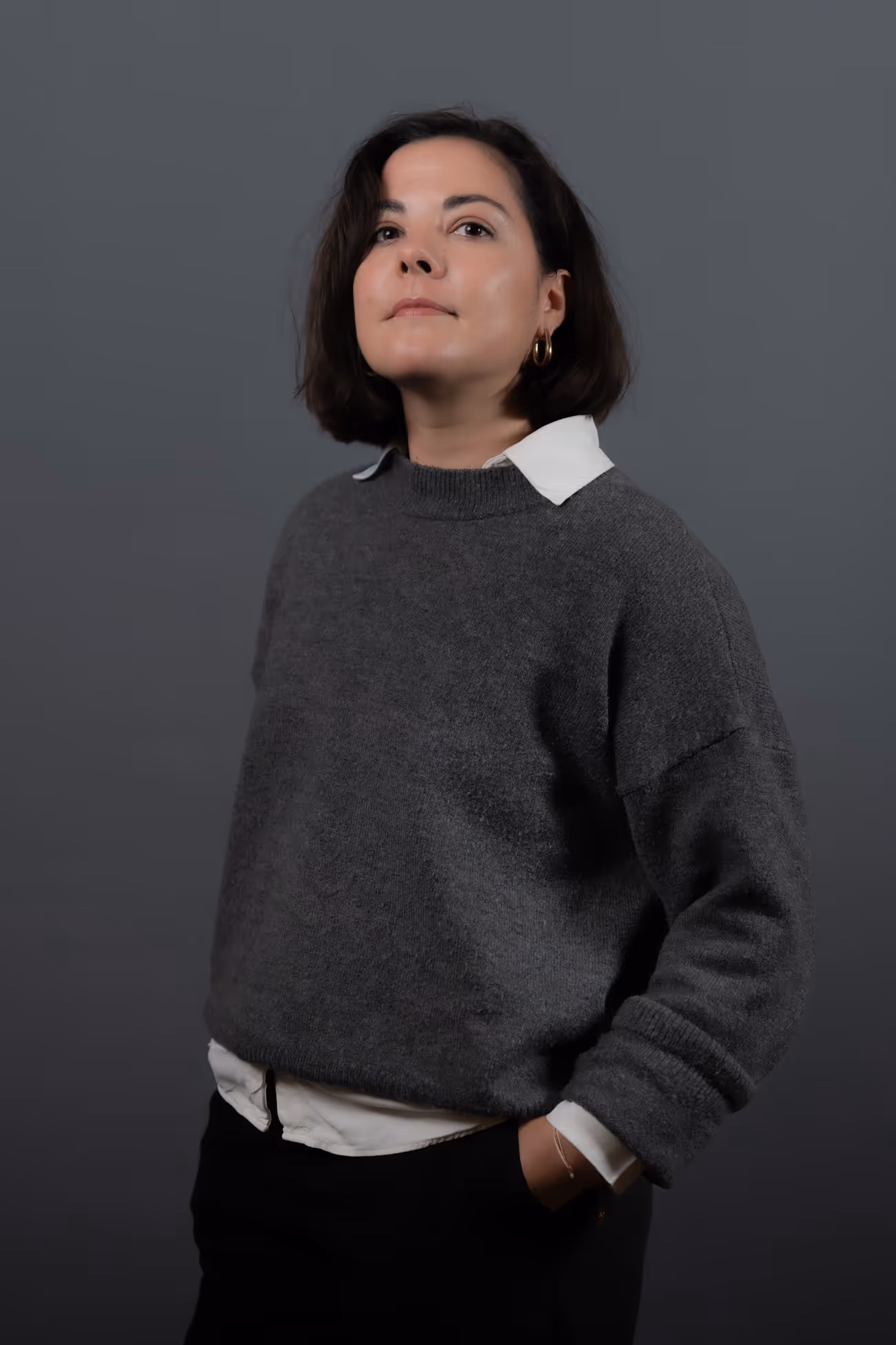 Portrait d'une femme aux cheveux courts portant un pull gris par-dessus une chemise blanche, posant avec la main dans la poche sur un fond gris.
