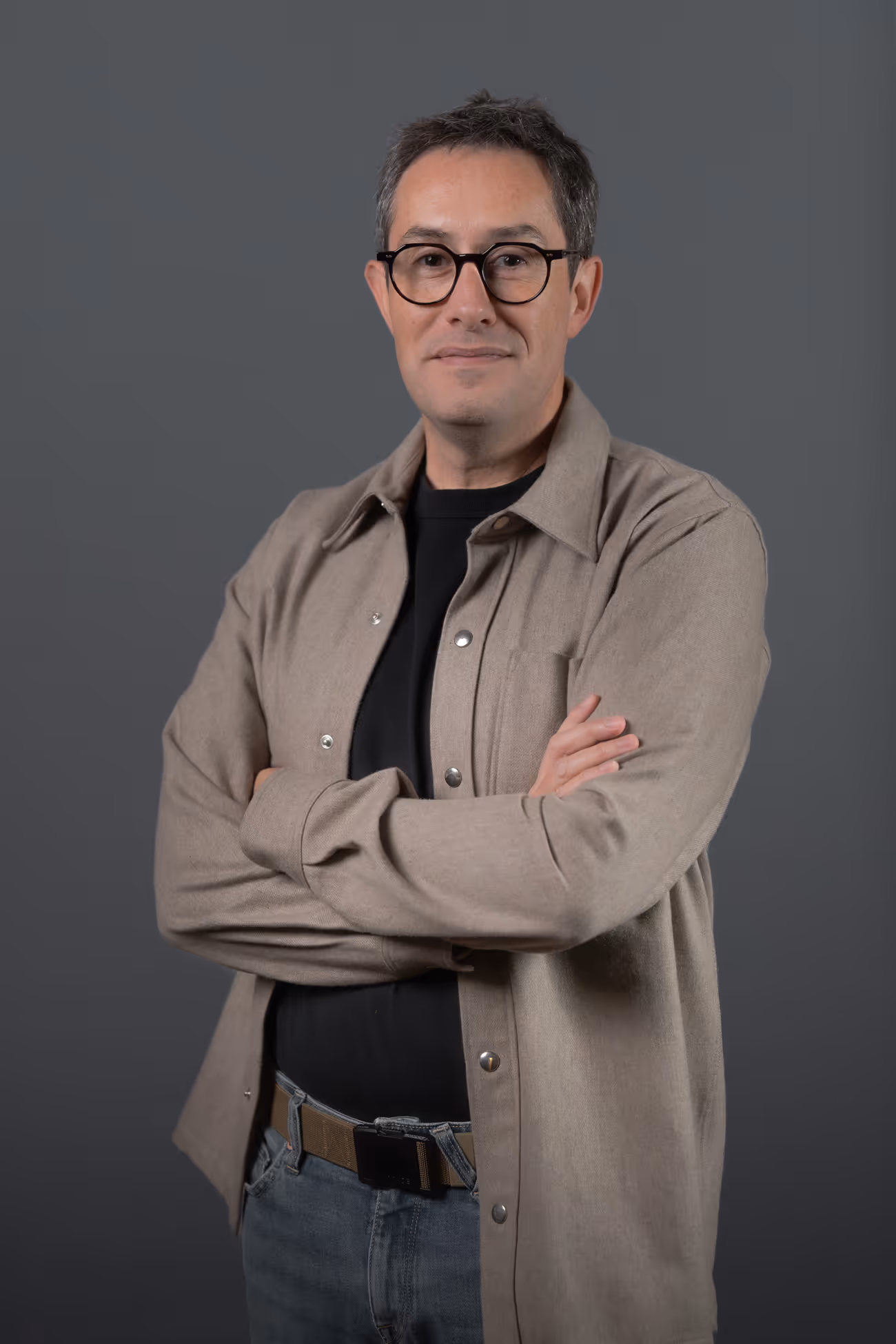 Homme portant des lunettes noires, veste beige et jeans bleus, bras croisés devant un fond gris.