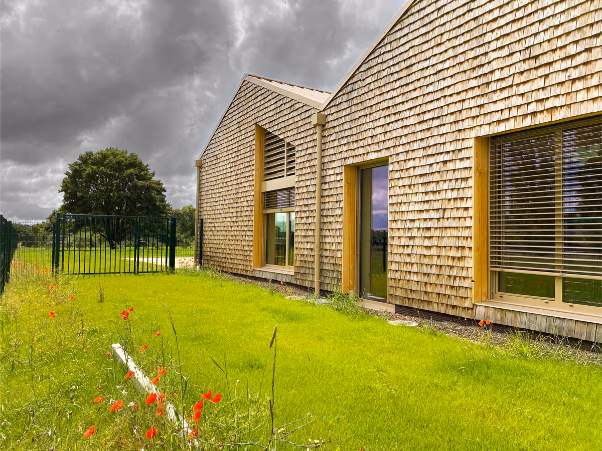 Maison moderne avec façade en bardeaux de bois, pelouse verte et fleurs rouges sous un ciel nuageux.