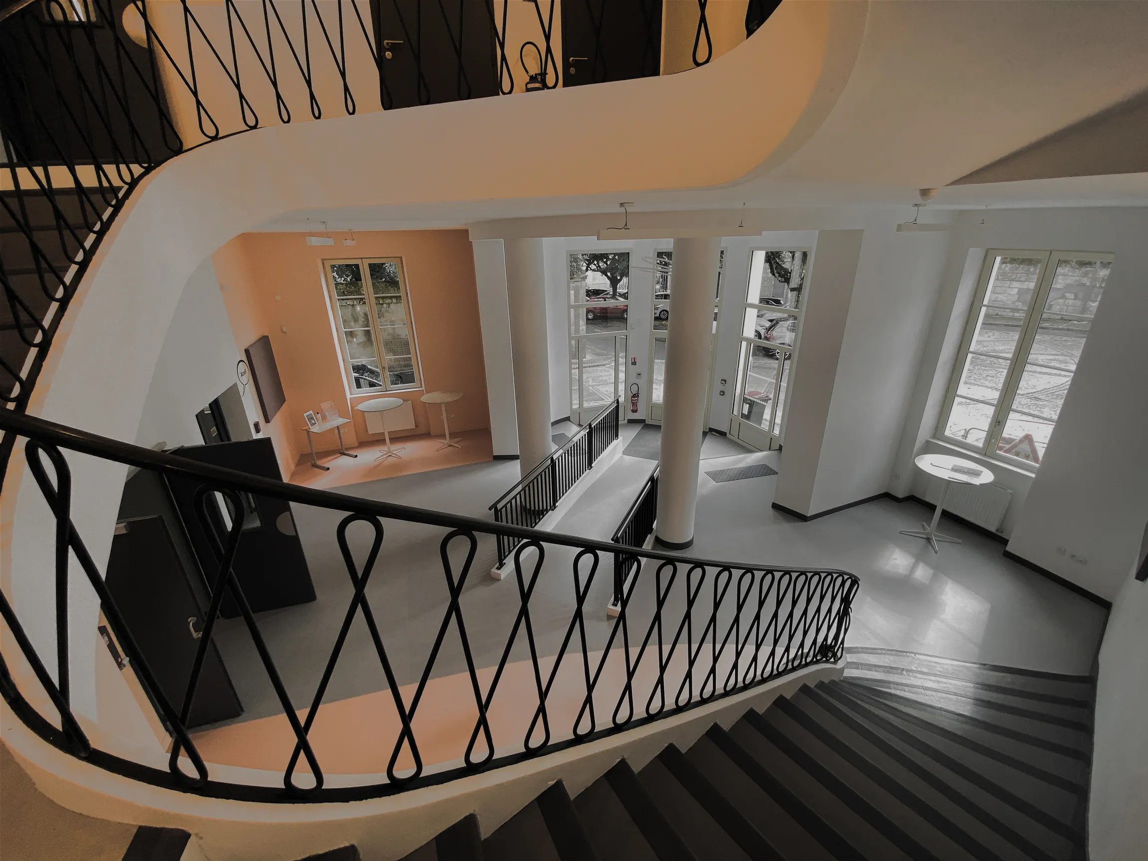 Intérieur d'un bâtiment avec un escalier en colimaçon, deux tables rondes et de grandes fenêtres laissant entrer la lumière naturelle.