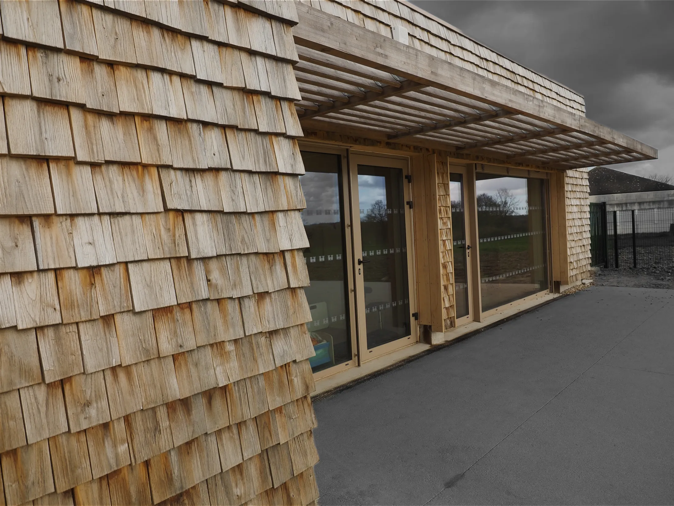 Façade en bois avec volets et grandes portes vitrées sous une pergola en bois.