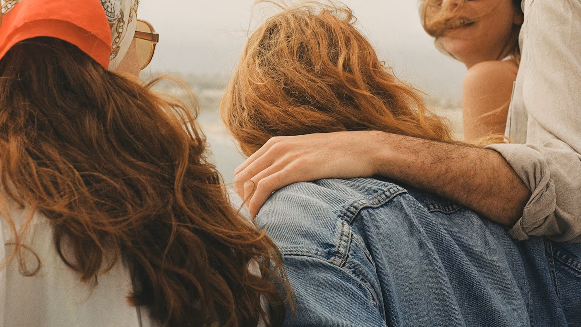 Back view of three people with long hair, one wearing a denim jacket, and a person’s arm resting on a shoulder.