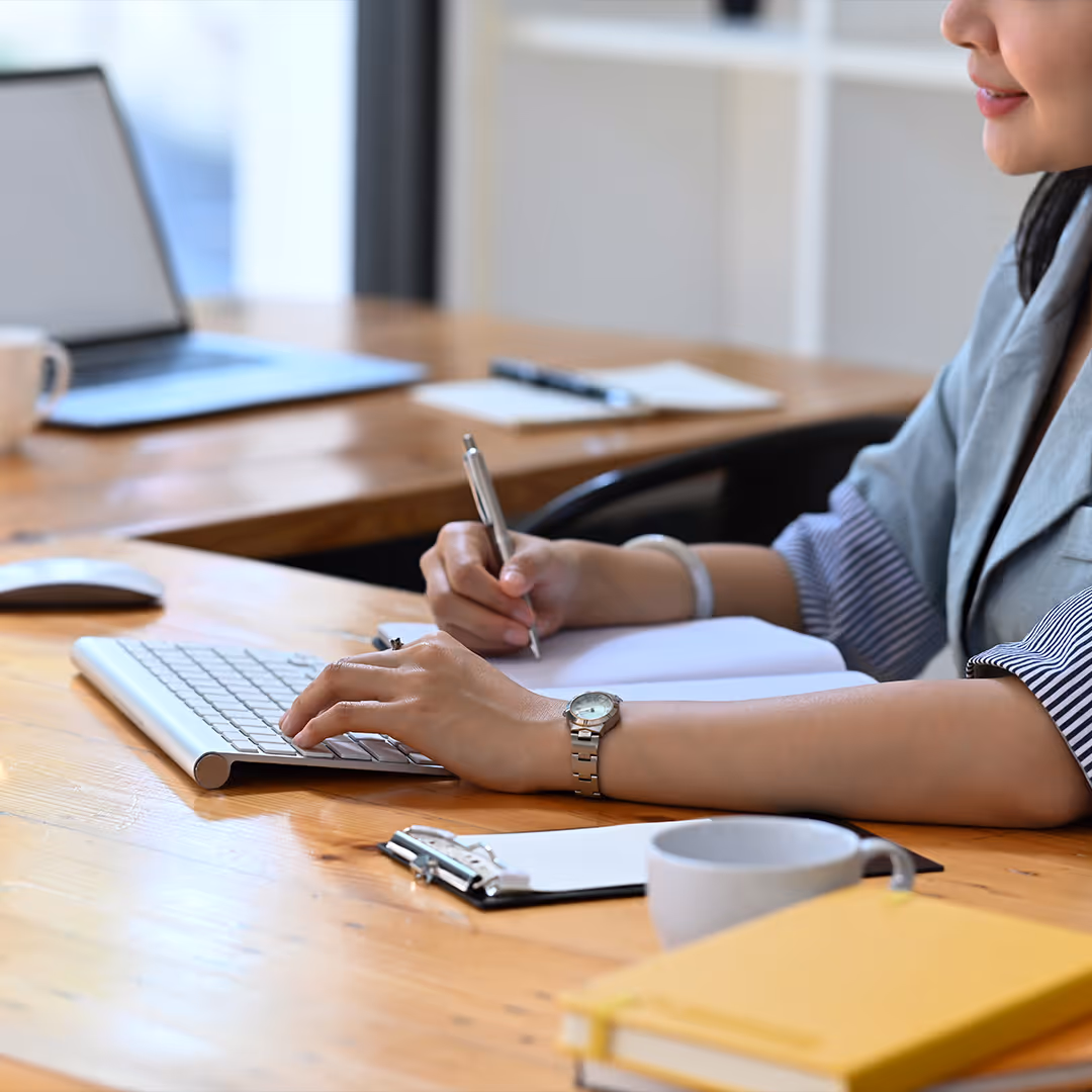 Person writing in a notebook while typing on a wireless keyboard at a wooden desk with a laptop and a coffee cup.