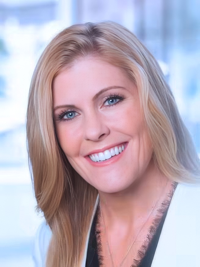 Smiling blonde woman with blue eyes wearing a white jacket in a bright indoor setting.