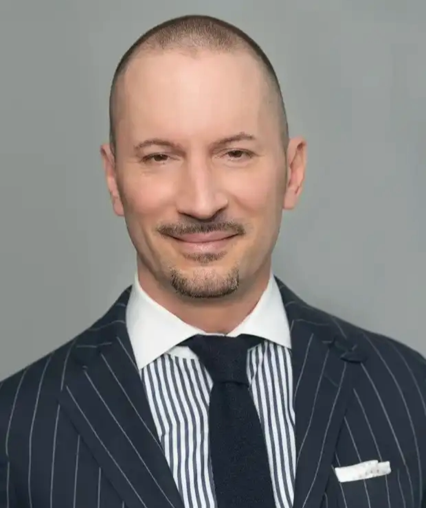 Smiling bald man with goatee wearing a navy pinstripe suit, striped shirt, and dark tie against gray backdrop.