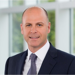 Bald man wearing a dark suit and patterned tie smiling in front of a windowed office background.
