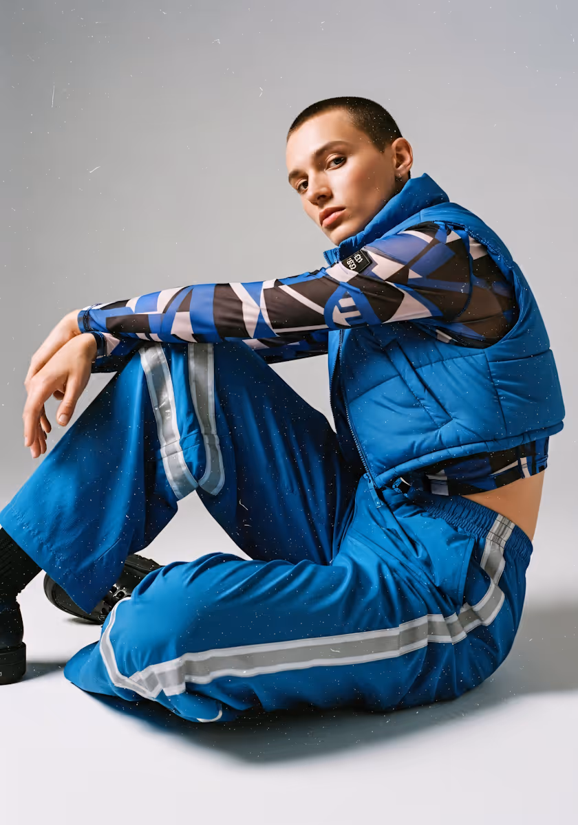 Person with short hair wearing a blue patterned long-sleeve top, blue puffer vest, and blue pants with reflective stripes, sitting on the floor.