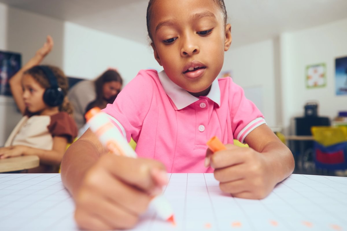 Young girl in a pink polo shirt concentrating on writing or coloring on a large grid paper at a classroom desk, with other children and an adult blurred in the background.