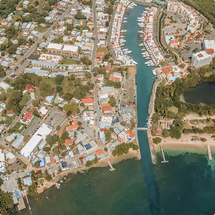 Aerial view of a coastal town with residential buildings, a marina with boats, canal, and green surroundings adjacent to a beach.